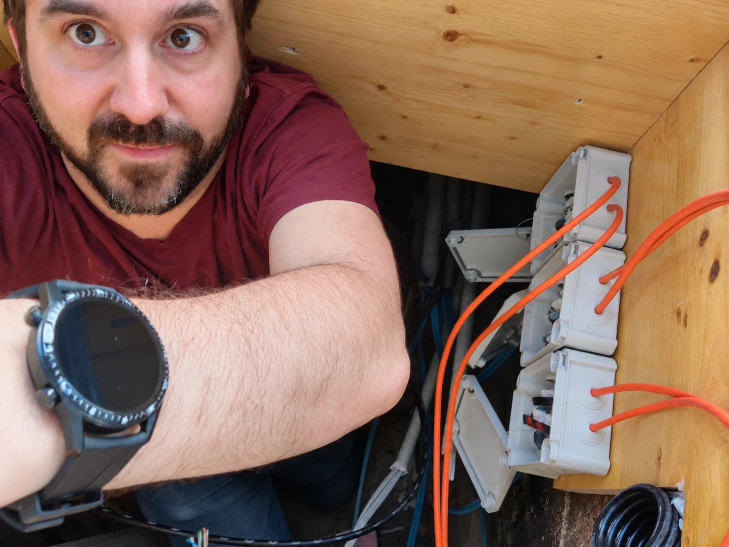Stefan Mayer in einem engen Schacht unter einem Holzboden, daneben Verteilerdosen mit orangefarbenen Kabeln.